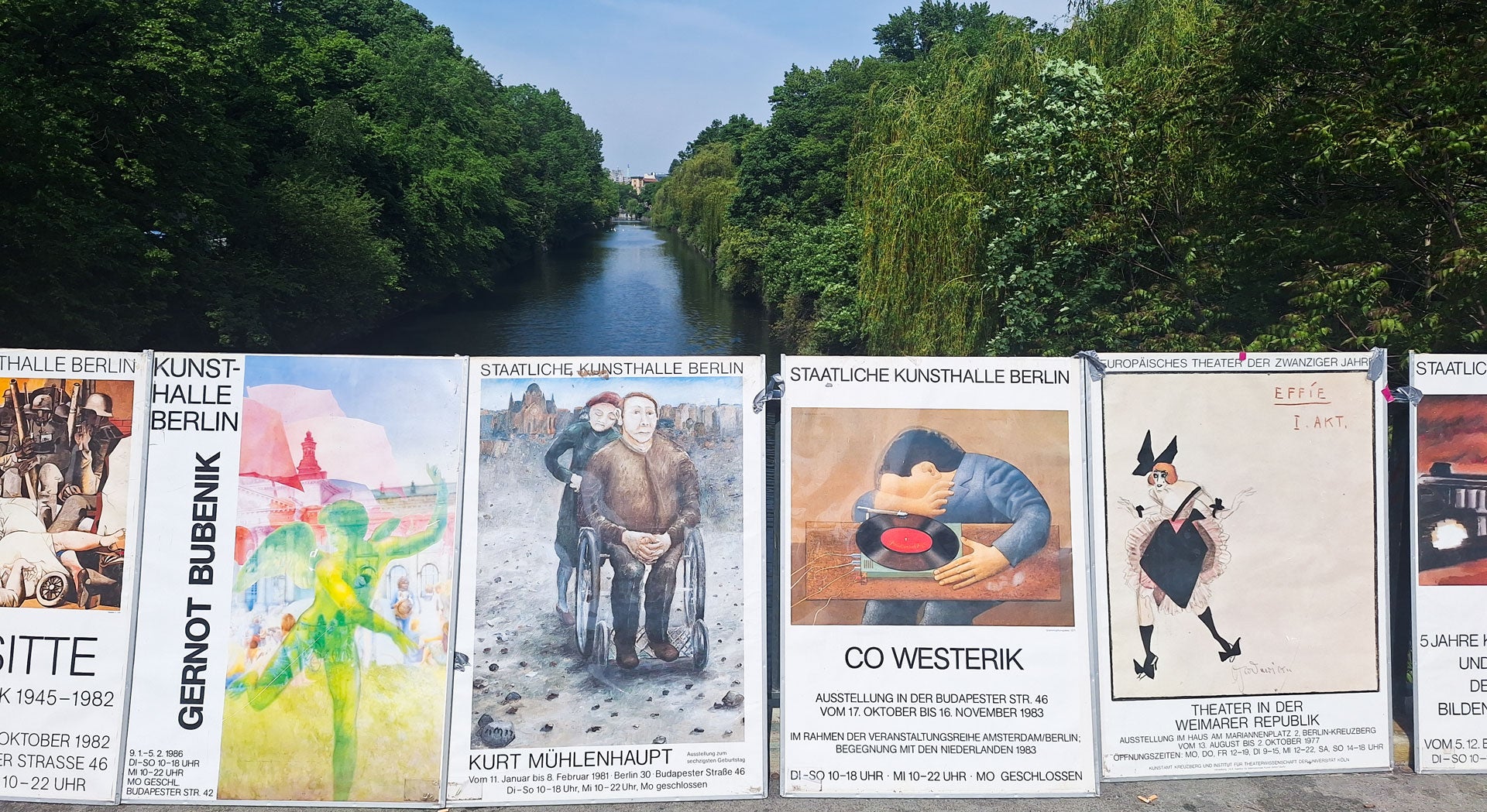 Verschiedene künstlerische Plakate im Vordergrund, verlinkt zu Produktseiten, mit einem Fluss und grünen Bäumen im Hintergrund